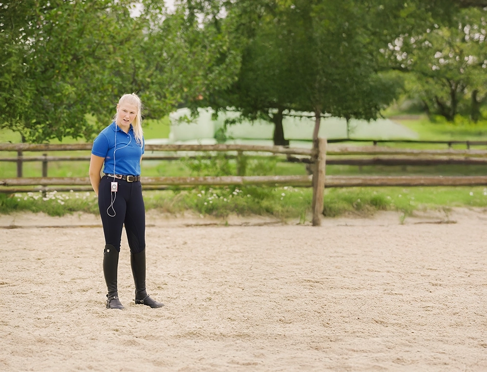 Reitlehrerin mit Kophörern und verschränkten Armen hinter dem Rücken auf Reitplatz.