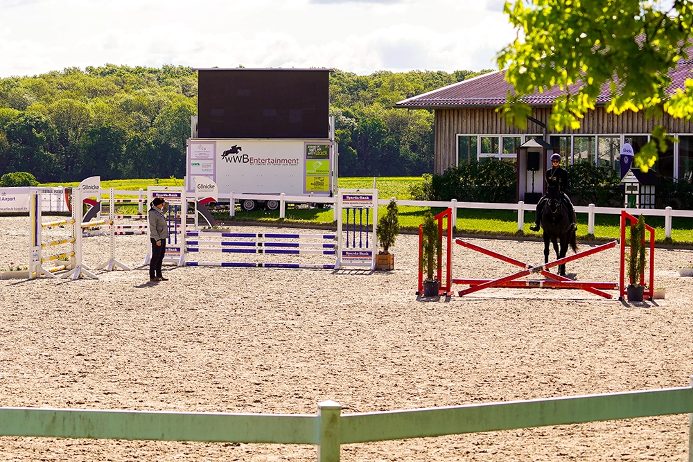 Reiterin auf schwarzbraunem Pferd überspringt Hindernis auf Reitplatz. Christina Pavel beobachtet.